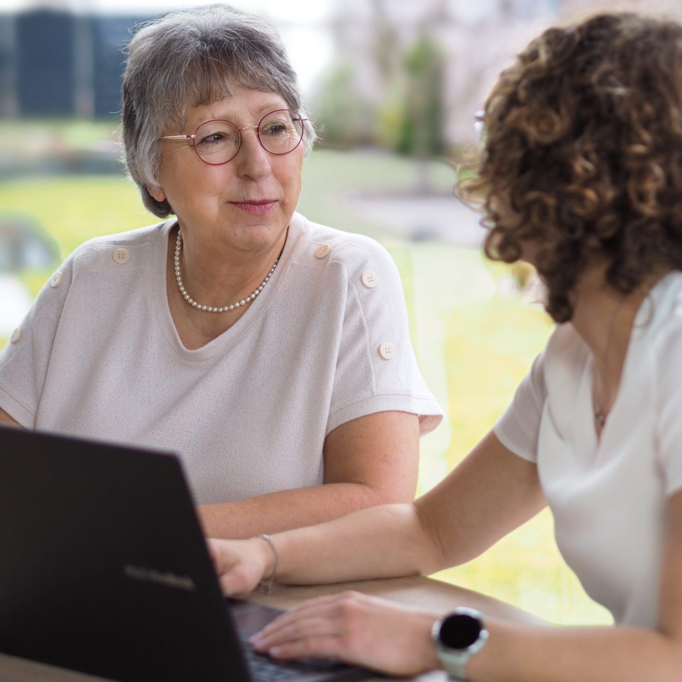 Zwei Frauen sitzen an einem Tisch, eine spricht, während die andere am Laptop arbeitet.